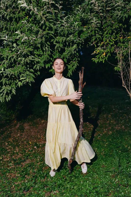 Woman in a flowing yellow dress pauses outdoors at night, holding a swing under trees, capturing a serene portrait in a quiet garden setting.