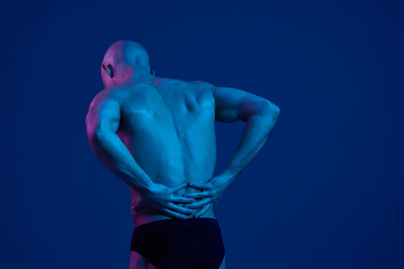 A man in a black and white image experiencing discomfort, highlighting a backache. The focus is on his muscular form against a vibrant blue background.