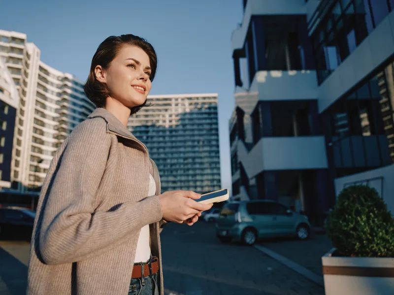 A cheerful professional woman stands outdoors in a modern urban setting, texting on a smartphone with a confident smile, creating a bright, dynamic business moment.