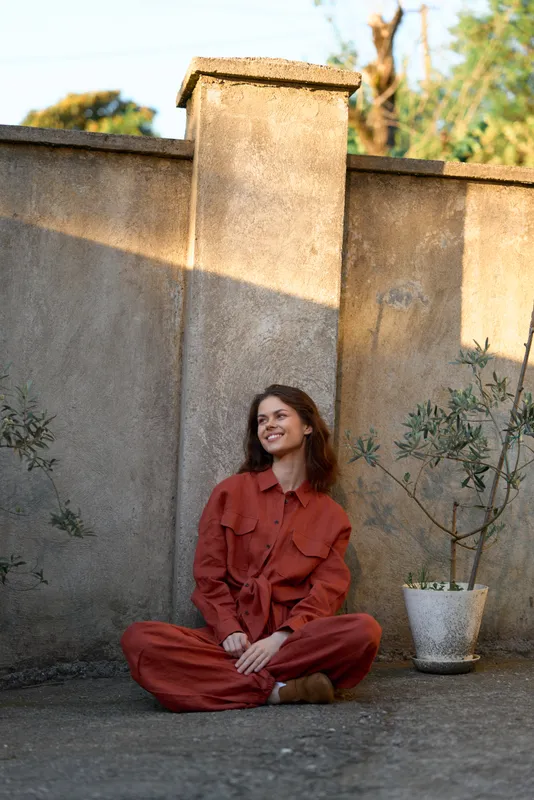 Young woman in a rust red jumpsuit sits cross legged on a rooftop terrace, smiling softly in warm afternoon light, relaxed against a concrete wall with a potted plant nearby