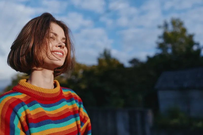 Woman smiling in a colorful striped sweater with short hair, young adult model in an outdoor sunlit countryside backyard. Joyful expression, casual outfit and lifestyle portrait for fashion and
