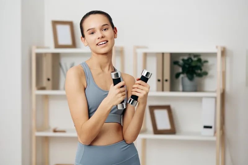 Athletic woman in sporty attire lifts dumbbells indoors, smiling during a workout. Clean studio backdrop with shelves, plants and framed decor, showcasing fitness, health and strength training.