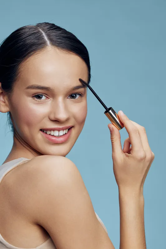 Young woman applying mascara for a beauty look, showcasing radiant skin and a cheerful expression against a light blue background