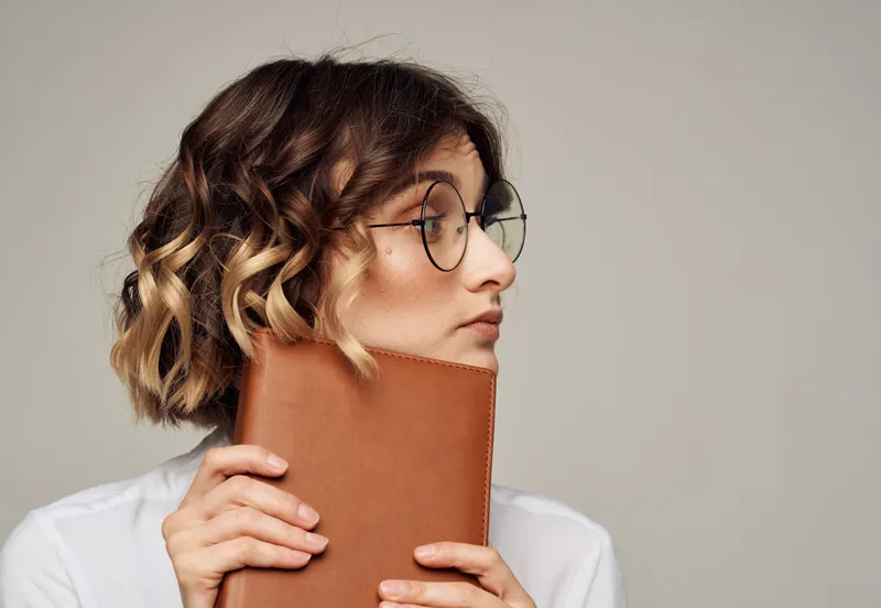 Романтичная женщина в очках на сером фоне держит в руках коричневую книгу