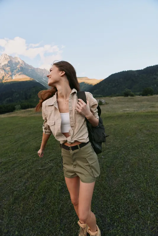 Cheerful woman outdoors, hiking in a sunny field near mountains, casual outfit with a backpack, light breeze, vibrant greens, sense of freedom and exploration