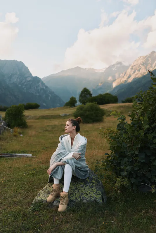 Woman sitting in a meadow with mountains and landscape in the background, outdoor fashion vibe, nature setting, calm relaxation mood, cozy outerwear.