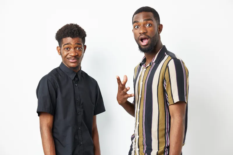 Two black men express surprise and joy with their playful poses against a neutral background. Their stylish outfits reflect contemporary fashion trends and energy.
