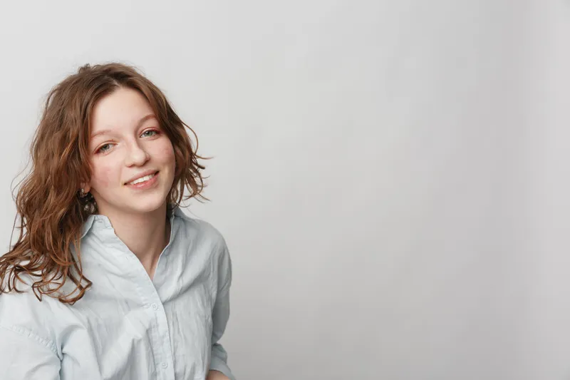 Smiling young woman with curly hair wearing a white shirt looks at camera on light grey background. Portrait of casual female model with natural expression and relaxed mood.