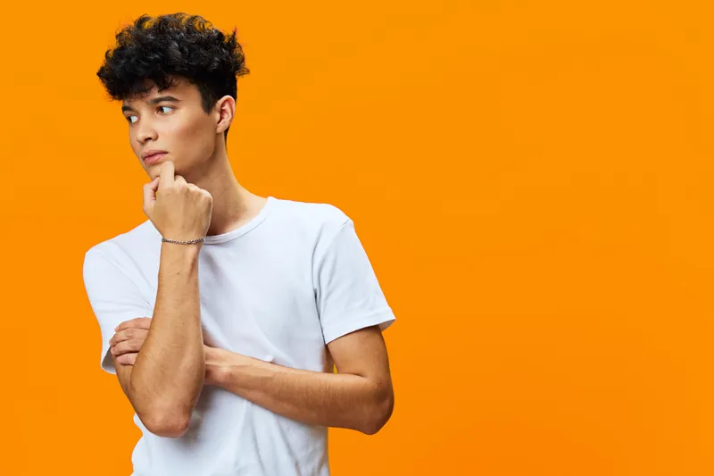 thoughtful man in white t-shirt with curly hair stands against vibrant orange background, looking sideways with pensive expression and casual posture