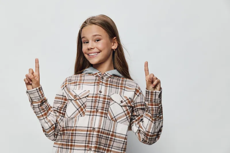 Girl smiling and pointing up with both hands wearing plaid shirt on white background happy child with long hair and casual style posing for camera.