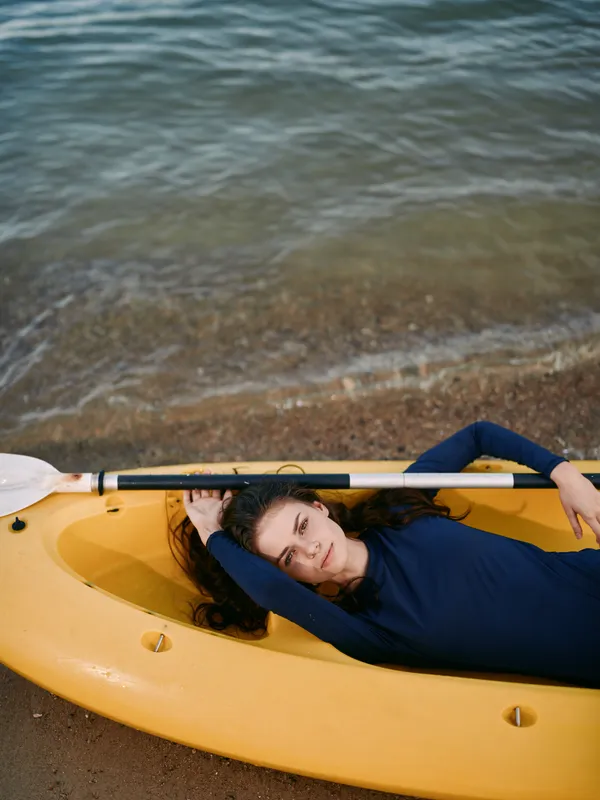 Relaxed woman lying in a yellow kayak on the beach near calm water with a paddle, enjoying peaceful outdoor recreation and leisure by the shore.