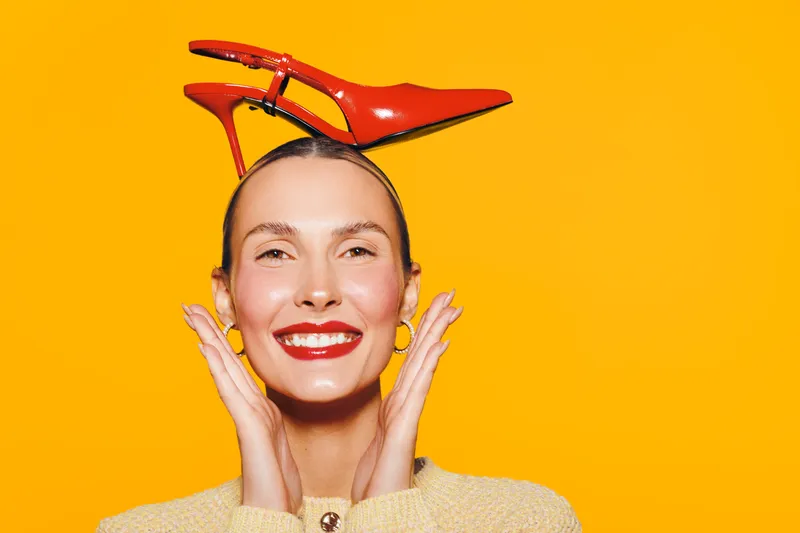 Smiling woman with red lipstick and yellow earrings holds red high heel shoe on her head against bright orange background. Concept of fashion, fun, and vibrant style.