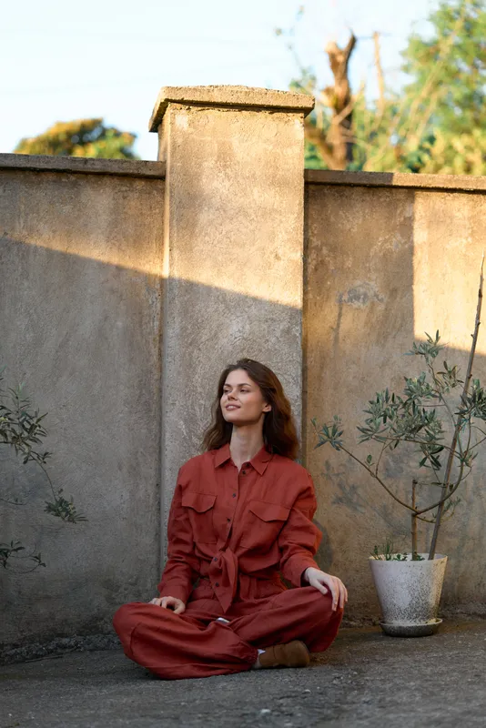 Woman sits cross legged in a red jumpsuit on a sunlit outdoor terrace, calm lifestyle portrait with rustic wall, potted tree, warm shadows, candid relaxed mood