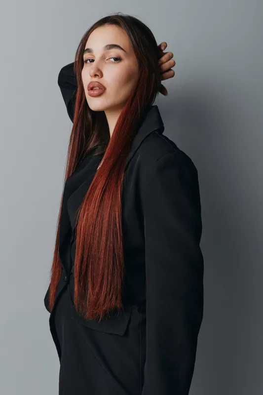 Young woman with long straight hair and stylish black blazer posing confidently against neutral background. Fashion portrait with natural makeup and casual elegance in studio setting.