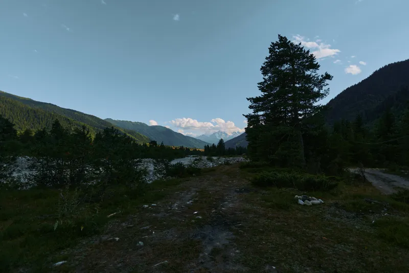 A tranquil mountain landscape unfolds under a clear blue sky. A winding dirt path leads toward a serene lake surrounded by evergreen trees and rugged hills.
