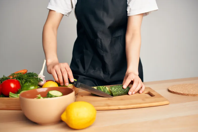 Женщина в черном фартуке нарезает овощи на кухне, домохозяйка готовит еду
