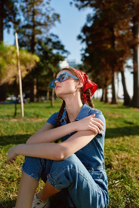 Lifestyle woman in modern boho-western denim street style wearing blue sunglasses and red bandana, sitting relaxed outdoors on green grass in warm film color light.