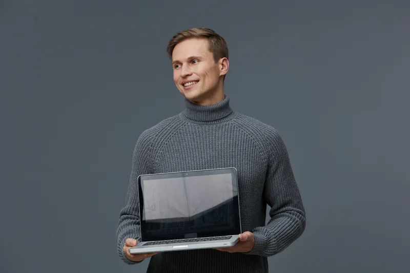Confident young man in gray turtleneck sweater holding open laptop device smiling looking away studio portrait isolated on plain gray background.
