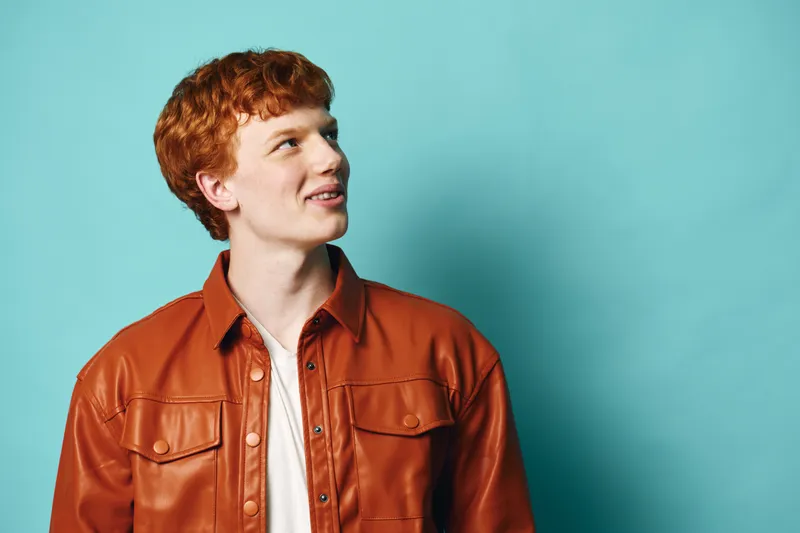 Young man with red hair wearing brown leather jacket smiling and looking up against blue background in studio portrait. Casual fashion, positive expression, isolated.
