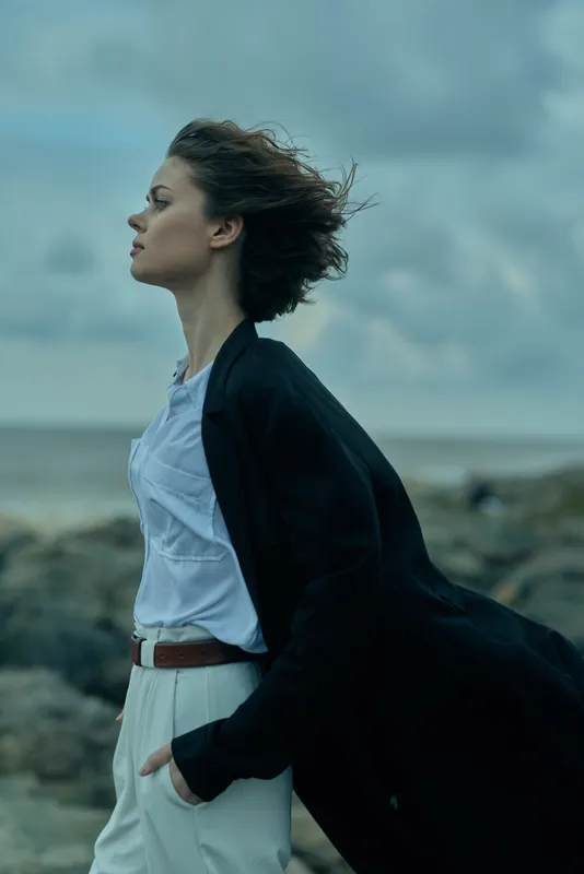 Wind swept profile on a rocky shore, wearing a white blouse, belt and dark coat, embodying calm confidence against the sea breeze in motion