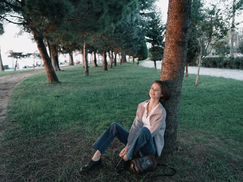 A relaxed individual sits against a tree in a park on a sunny day, smiling softly and enjoying a moment of calm, creating a peaceful outdoor atmosphere.