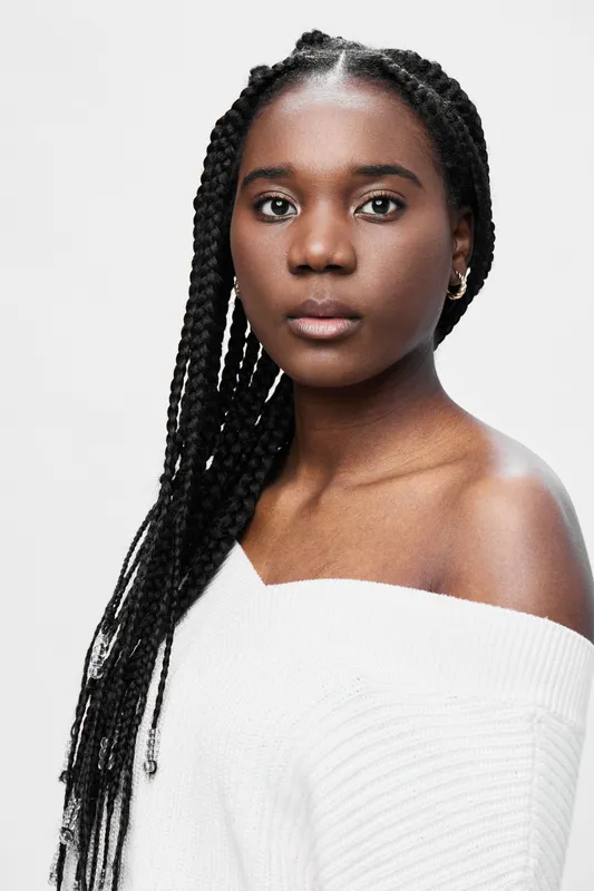 Portrait of a young Black woman with braided hair, wearing a white off shoulder sweater, conveying a sense of confidence and elegance against a neutral background