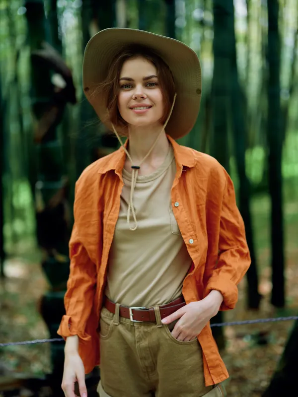 woman forest hat smile orange outdoors casual portrait of a young traveler in an orange shirt and khaki shorts, standing among trees, enjoying sunlight and nature with relaxed confident expression.