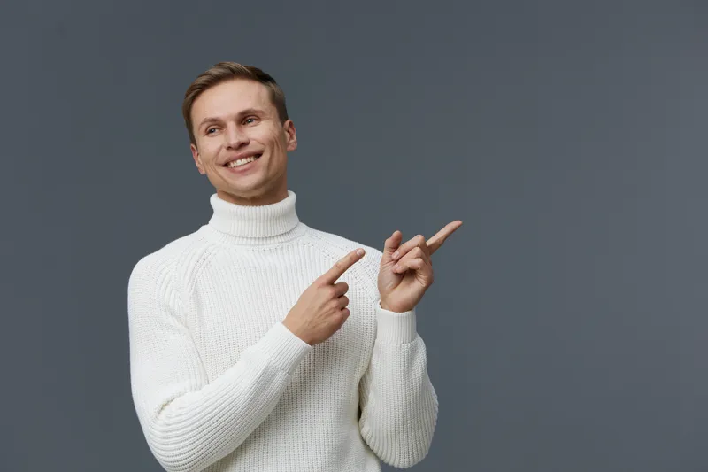 Bright smiling young man with short brown hair wearing a cozy white turtleneck sweater pointing to the right, looking cheerful, happy, and friendly, isolated on gray background, studio portrait. Peopl