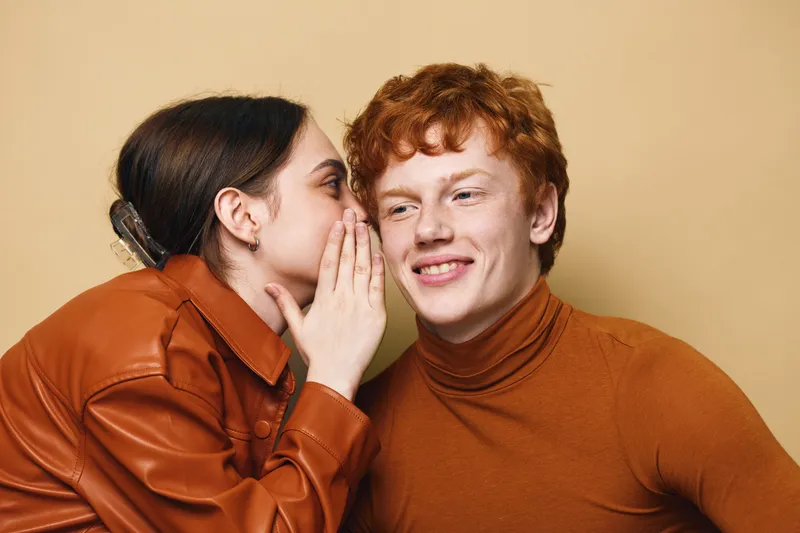 Young woman whispering secret to young man, closeup portrait with smile. Friendship, communication and intimacy with casual clothes on beige background in studio.