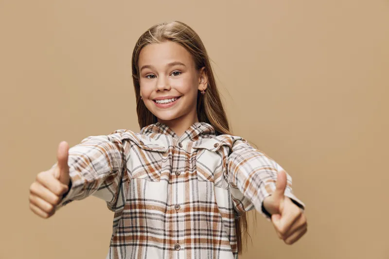 Girl in plaid shirt smiling and showing thumbs up gesture with both hands on beige background. Positive expression, casual clothing and long hair, kid portrait with happiness and confidence.