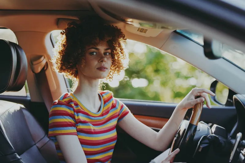 Young woman drives a stylish car with sunlight streaming through the window as her curly hair frames a calm confident expression and a striped tshirt accents color