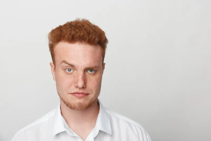 Portrait of a serious young man with red hair and green eyes wearing a white shirt. Neutral expression on a plain light background. Concept of confidence, youth, and natural appearance.