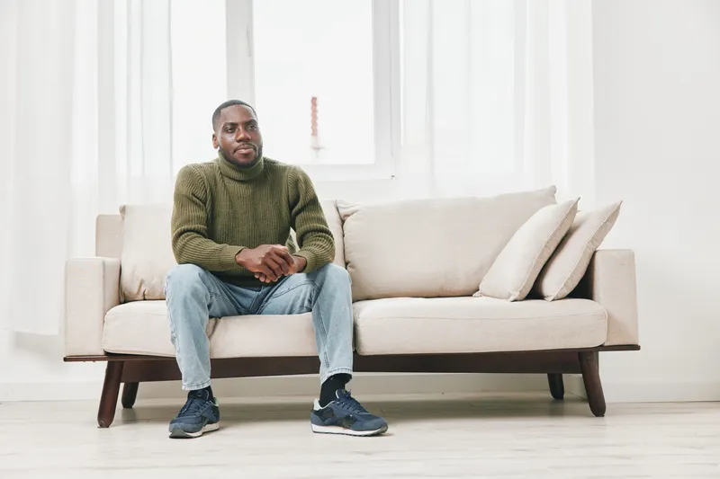 Relaxed African American man in a cozy sweater sitting on a modern sofa, conveying comfort and tranquility in a bright, minimalist living room