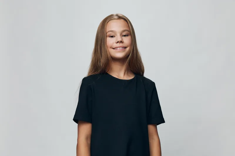 Girl, child and smile with long hair wearing black t shirt standing against gray background. Portrait, youth and happiness with casual clothing for childhood and beauty concept.