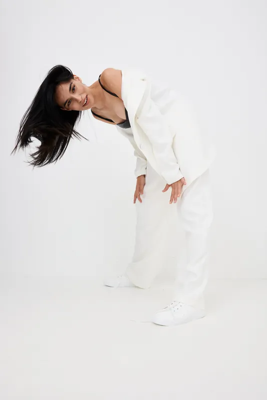 Asian woman in white suit bending forward with flowing hair, wearing black top and sneakers, against clean white background in stylish fashion pose.