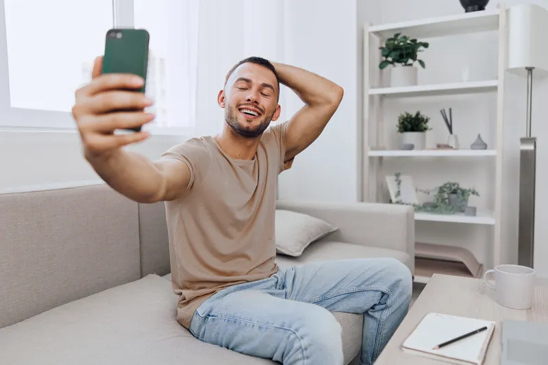 Happy man taking a selfie on a couch in a modern living room, showcasing the joy of casual moments and digital communication in 2025 style.