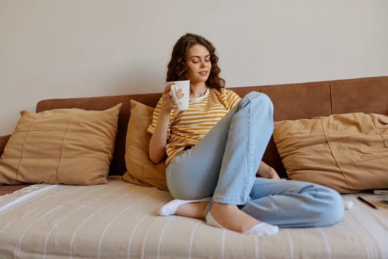 Casual woman at home relaxing on a sofa, holding a cup, striped shirt and jeans, soft natural light, cozy living room atmosphere, peaceful moment, everyday lifestyle