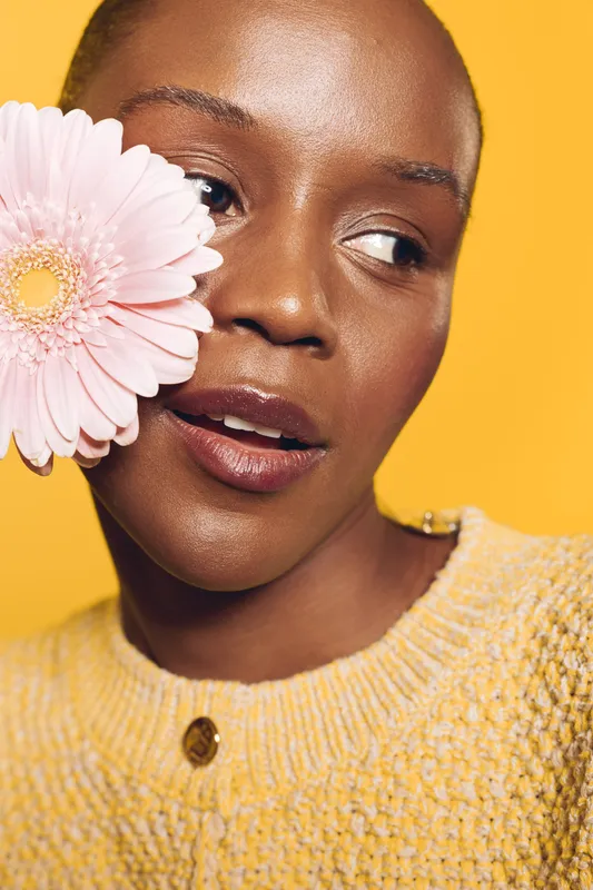 Close-up of a dark skinned African woman with natural makeup holding a pink flower near face. Yellow background and knitted sweater create warm, fresh concept of beauty and nature.