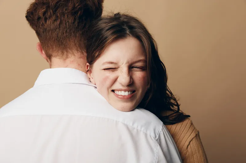 Happy young woman with closed eyes smiling while hugging man in white shirt. Intimate moment of affection and love between couple on beige background. Concept of relationship and joy.