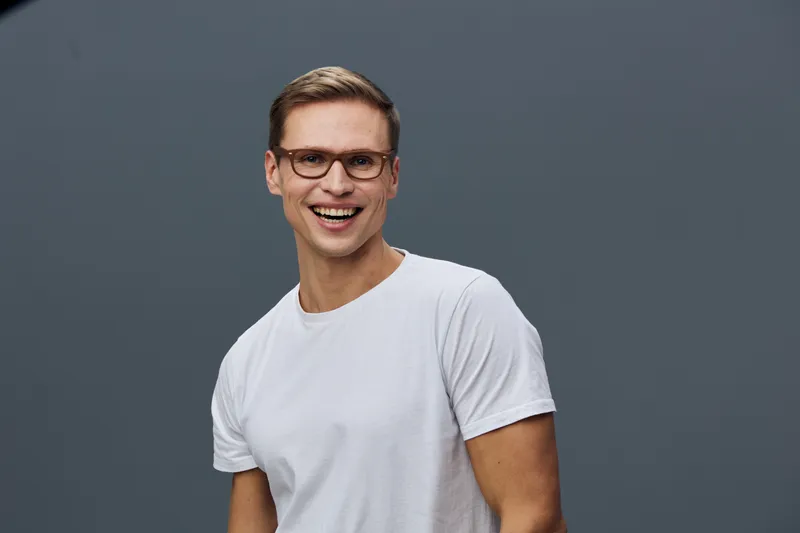 Smiling man wearing white t-shirt and glasses, looking cheerful and confident, studio portrait with plain gray background, casual fashion, positive expression, eye contact, friendly vibe, horizontal c