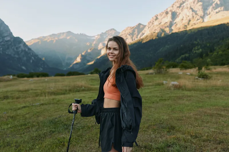 A cheerful female hiker stands in a grassy valley with a trekking pole, smiling at the camera as rugged mountains rise behind her under a clear blue sky.