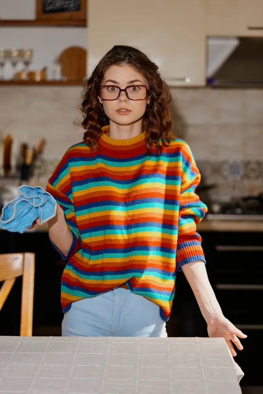 Young woman with curly hair and glasses wears a colorful striped sweater in a kitchen, wiping a surface with a cloth, capturing a candid moment of home life and concentration.