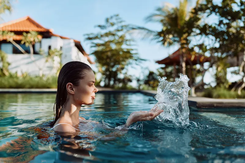 Woman in a pool creates a splash with her hand, serene expression, tropical resort setting, sunlit water, palm trees, lifestyle portrait of leisure and outdoor relaxation
