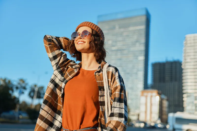 Woman smile city sunlight urban portrait casual beanie and sunglasses, candid lifestyle moment with authenticity and golden hour glow. Mindful living, emotional storytelling in modern street scene.