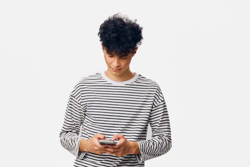 young man with curly hair wearing striped shirt looking down at smartphone with focused expression on white background, casual style and modern technology theme
