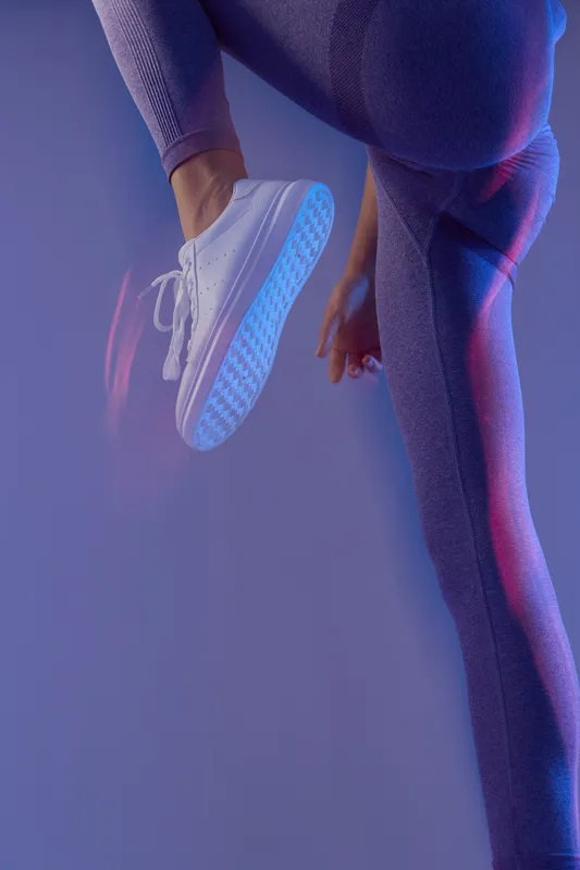 Woman sport fitness in stylish purple leggings and white sneakers performing dynamic movement against a soft blue and pink lit background, emphasizing health and agility.
