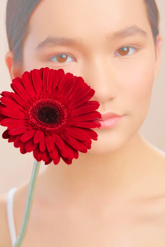 asian woman poses with a vibrant red flower close to her face, soft lighting and calm expression create an elegant, contemporary portrait with a minimal, dreamlike mood.