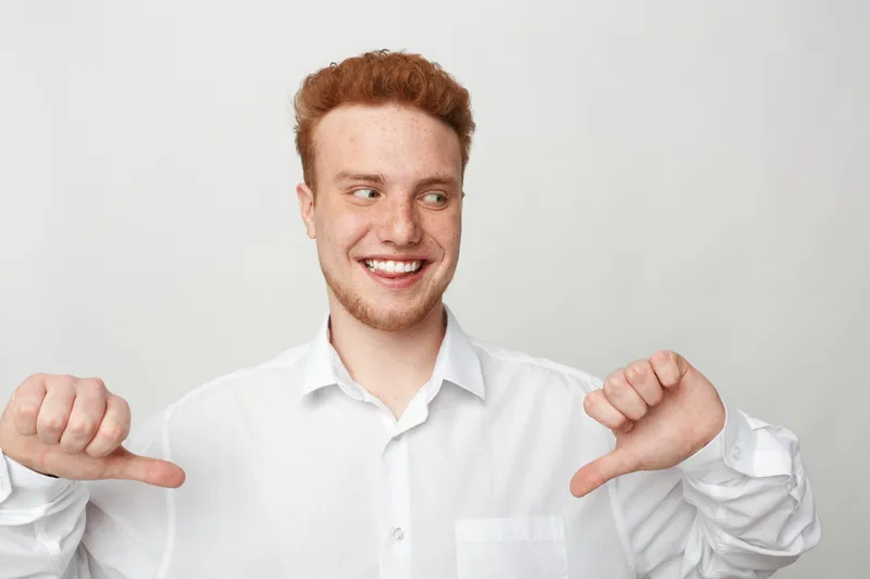 Smiling young man with red hair and beard wearing white shirt points thumbs at himself on light background. Concept of confidence, self pride, happiness, and positive attitude.