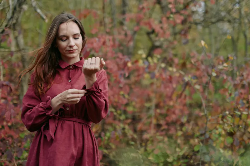 Authentic portrait of a thoughtful woman in a burgundy dress, capturing a genuine moment and honest mood amid an autumn forest setting with soft light.