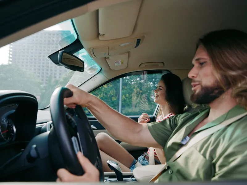 A candid moment inside a car shows a bearded man driving while a cheerful woman sits in the passenger area, enjoying the ride and sunny street views.